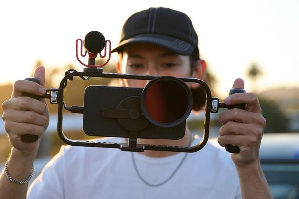 Hand holding iPhone 16 with a mobile filmmaker cage with an on-camera shotgun microphone for footage.